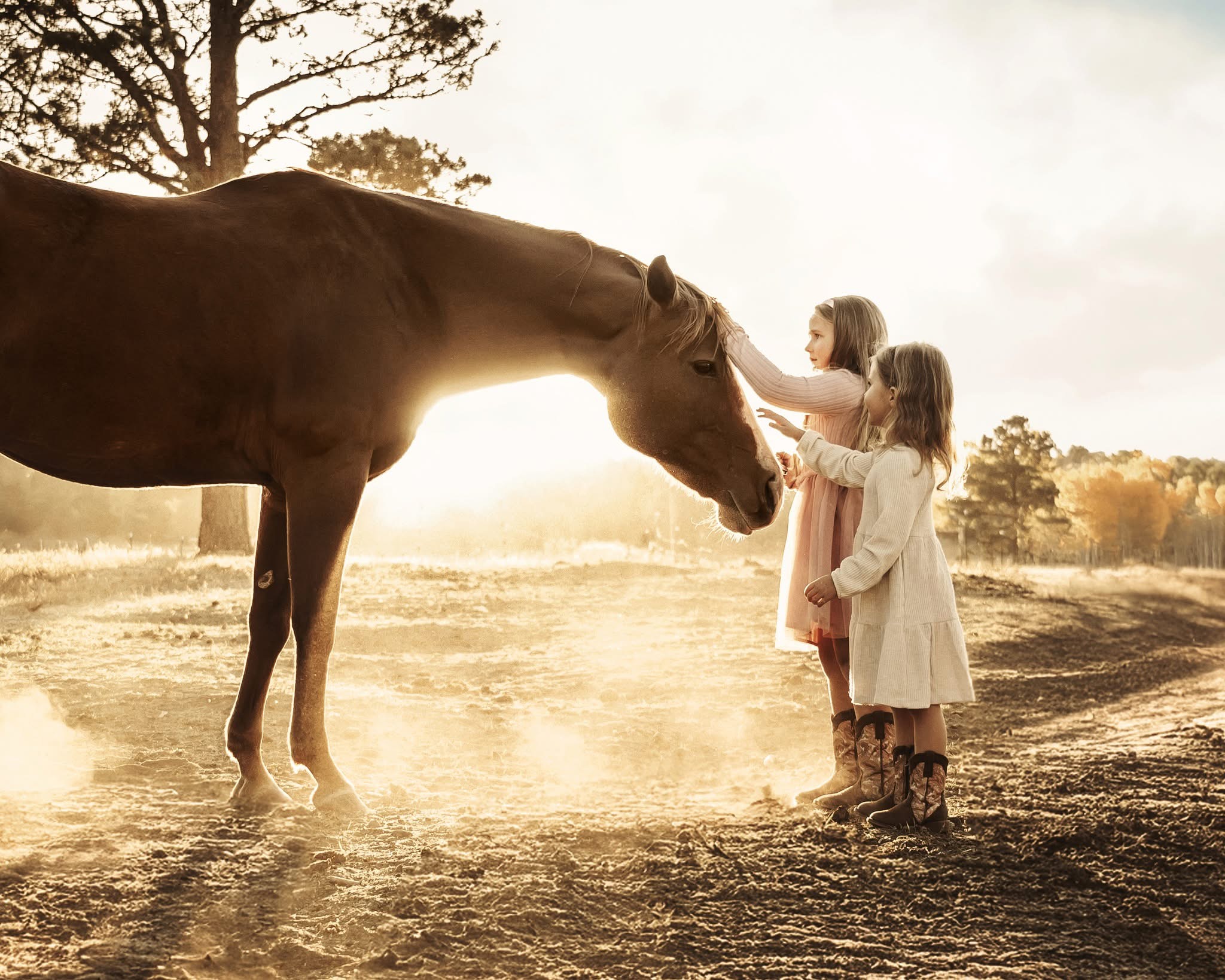 Two young girls stand on a dusty path, gently petting a brown horse at sunset with trees in the background.